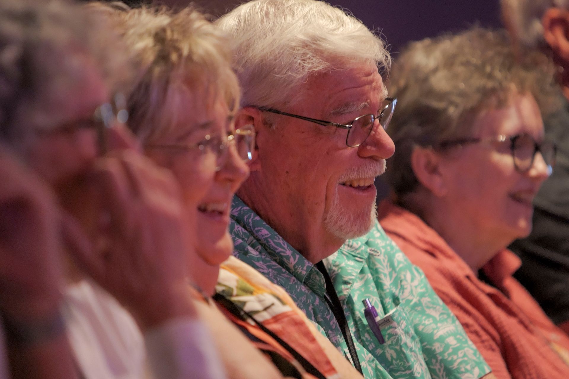 Group of smiling people, watching something. Man with glasses and light green shirt, others with glasses.