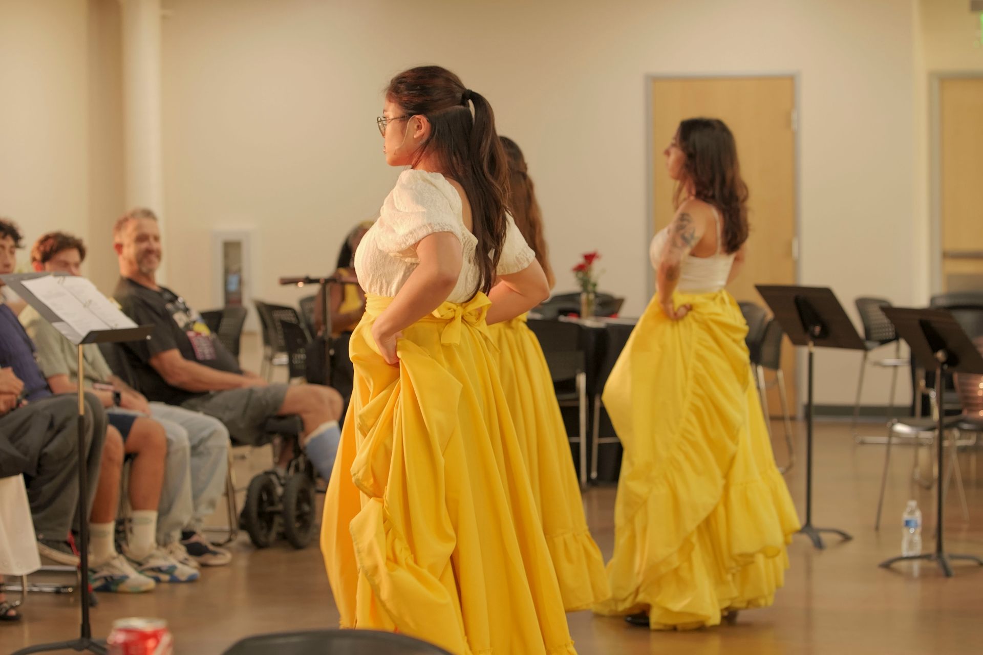 Two women in yellow skirts and white tops dance on a stage. Audience watches.