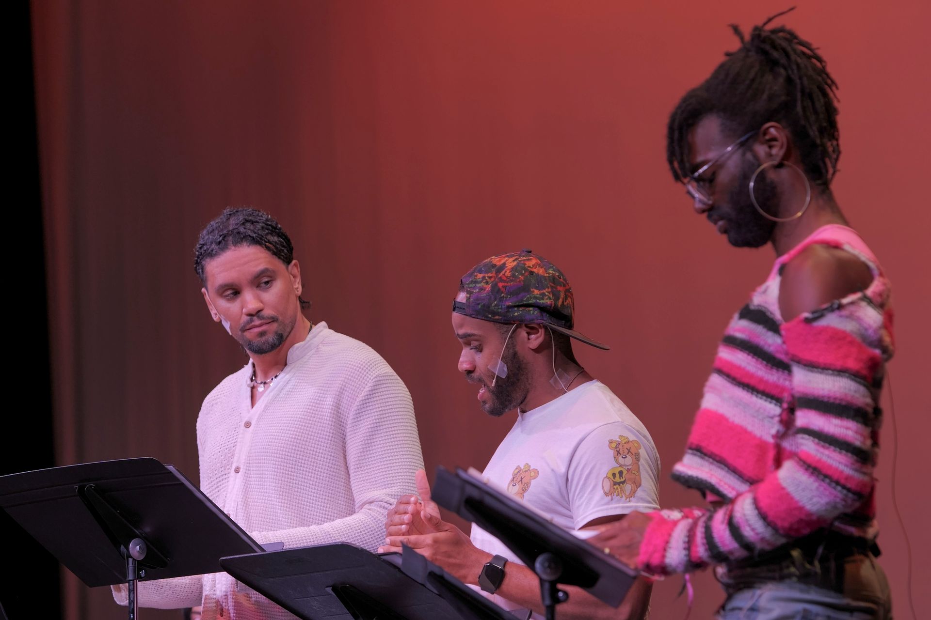 Three people reading from scripts. Two men on the left in a white tops. Third person wears glasses and pink-striped sweater.