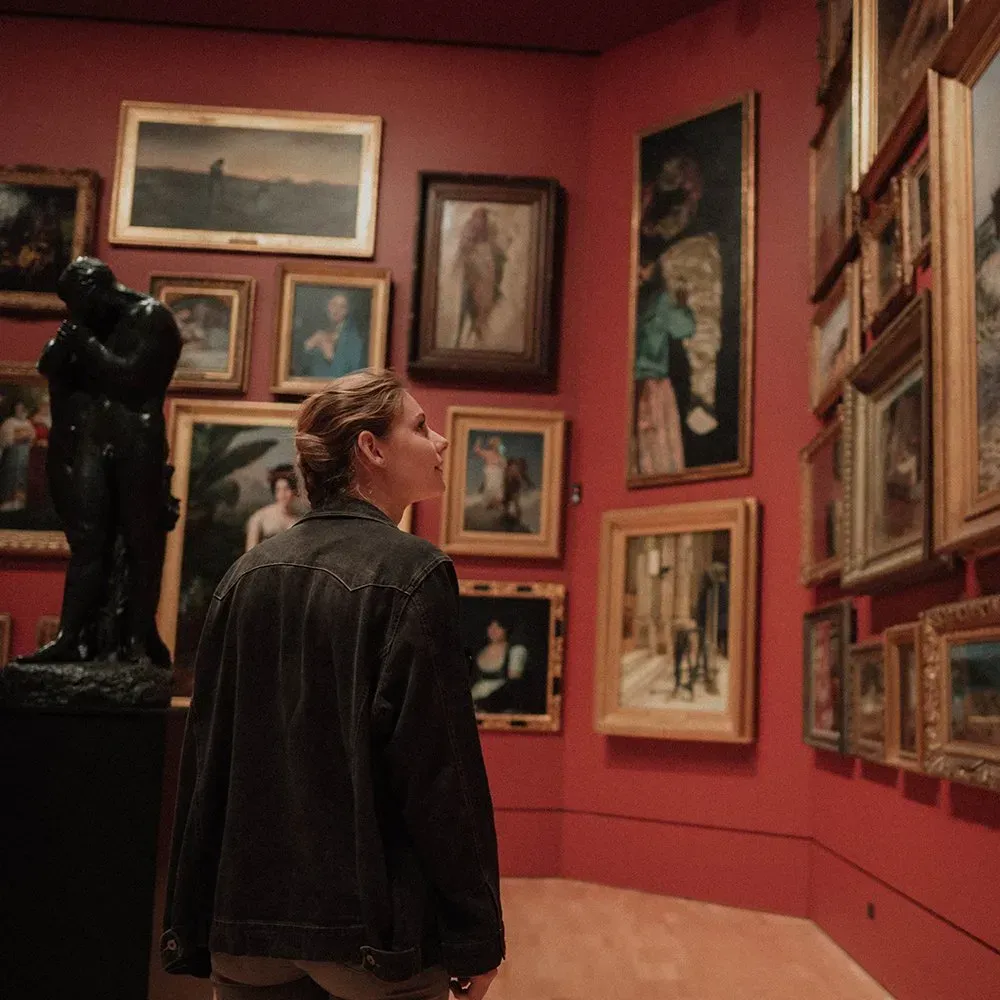 Person viewing paintings in a red-walled gallery. Many framed artworks are on display.