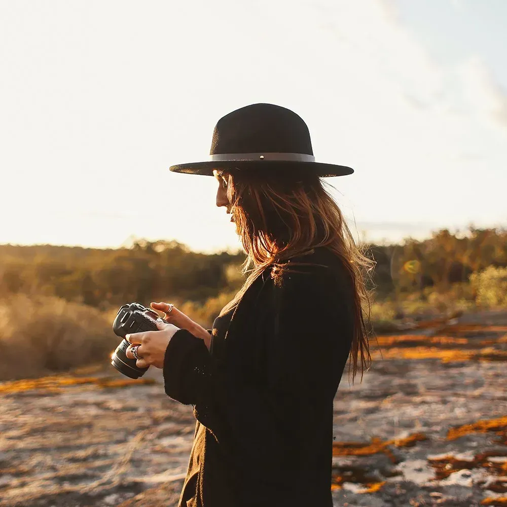 Woman in black hat and coat, holding a camera outdoors, looking at the camera.