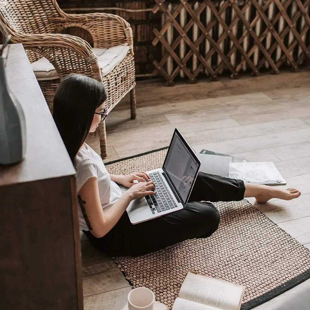 Woman sitting on rug, working on laptop, with book and coffee.