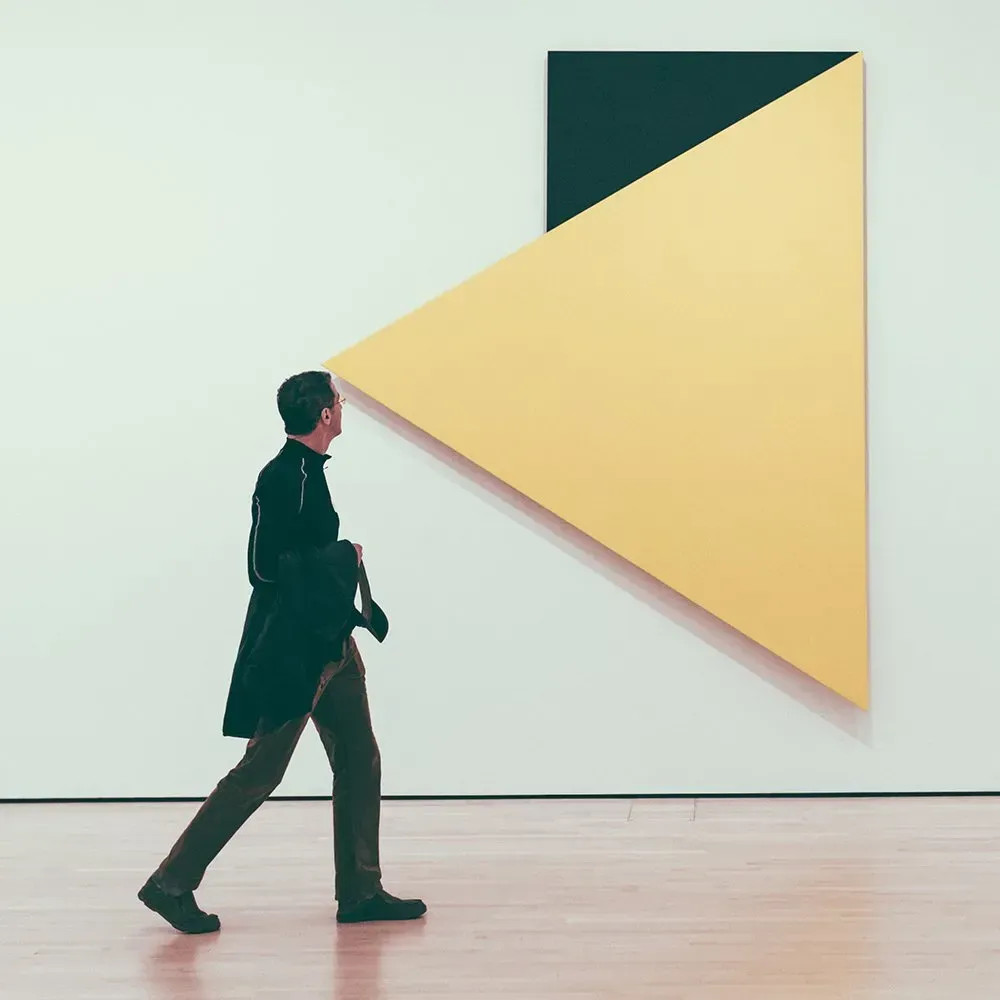 Man walks past a modern art painting on a white wall; yellow triangle and black rectangle.