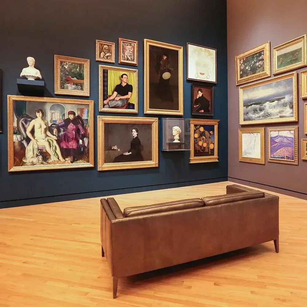Gallery room with many framed paintings on a blue wall. A brown couch sits in front. Wooden floor.