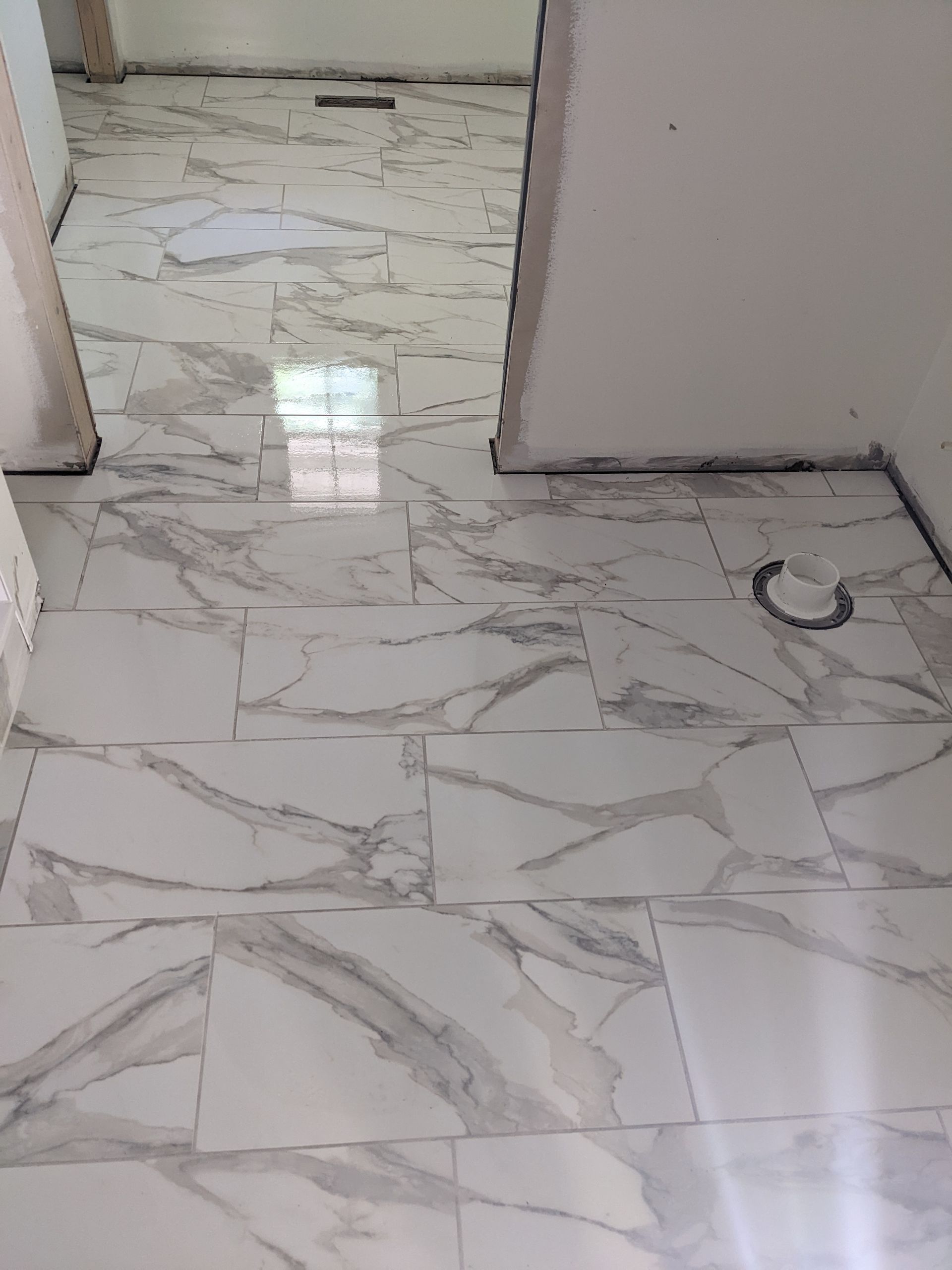 White and gray marble-patterned tile flooring in a room with white walls and doorways; a drain and plumbing visible.