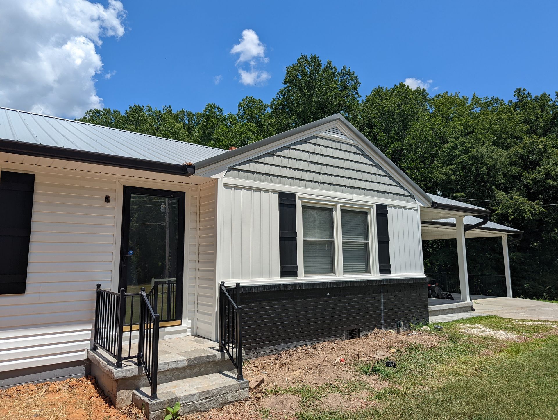 White and black house with a metal roof on a sunny day. Features a porch, shutters, and is surrounded by trees.