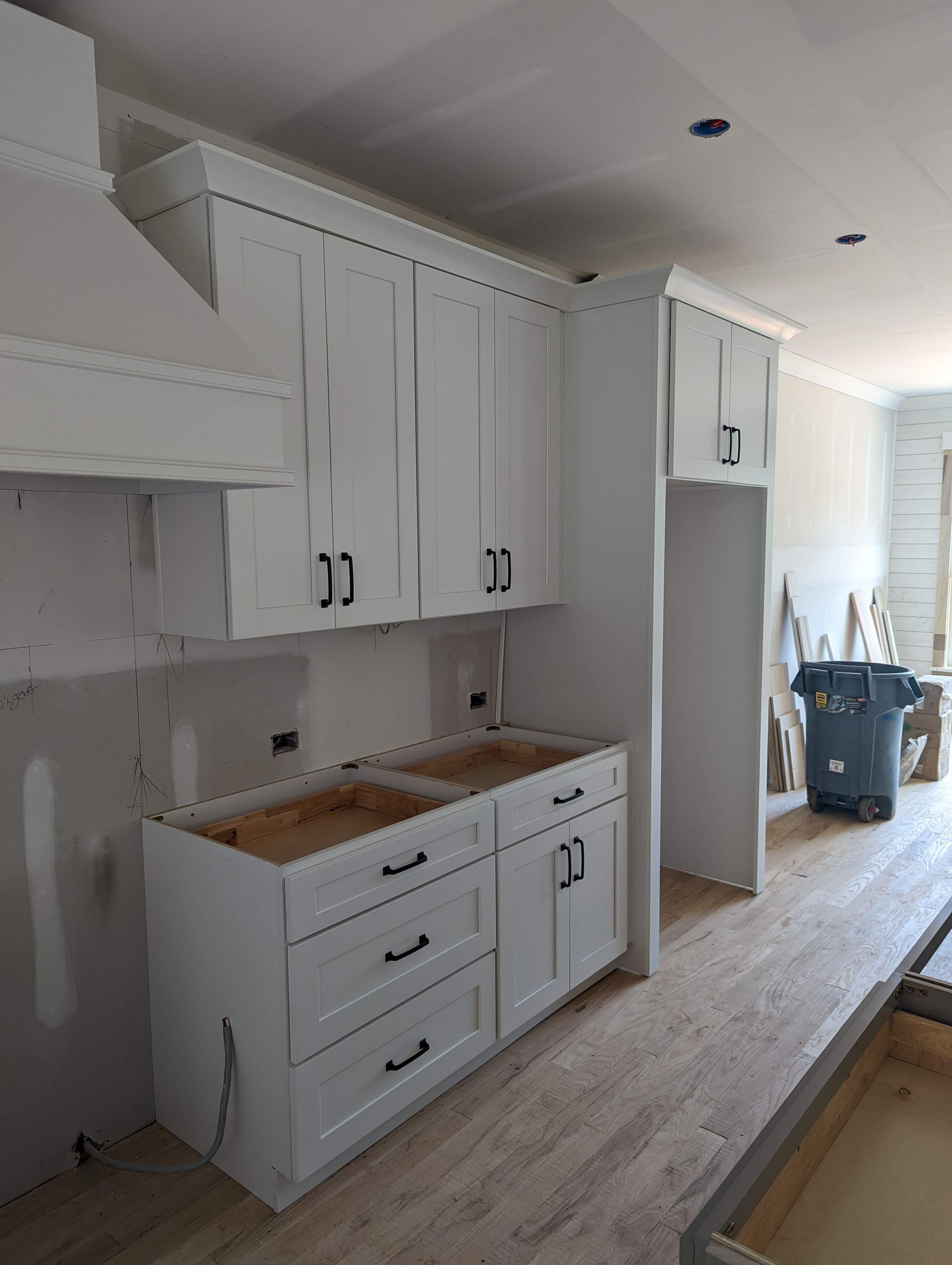 White kitchen cabinets with black hardware in a room under construction. A range hood and refrigerator enclosure are also present.