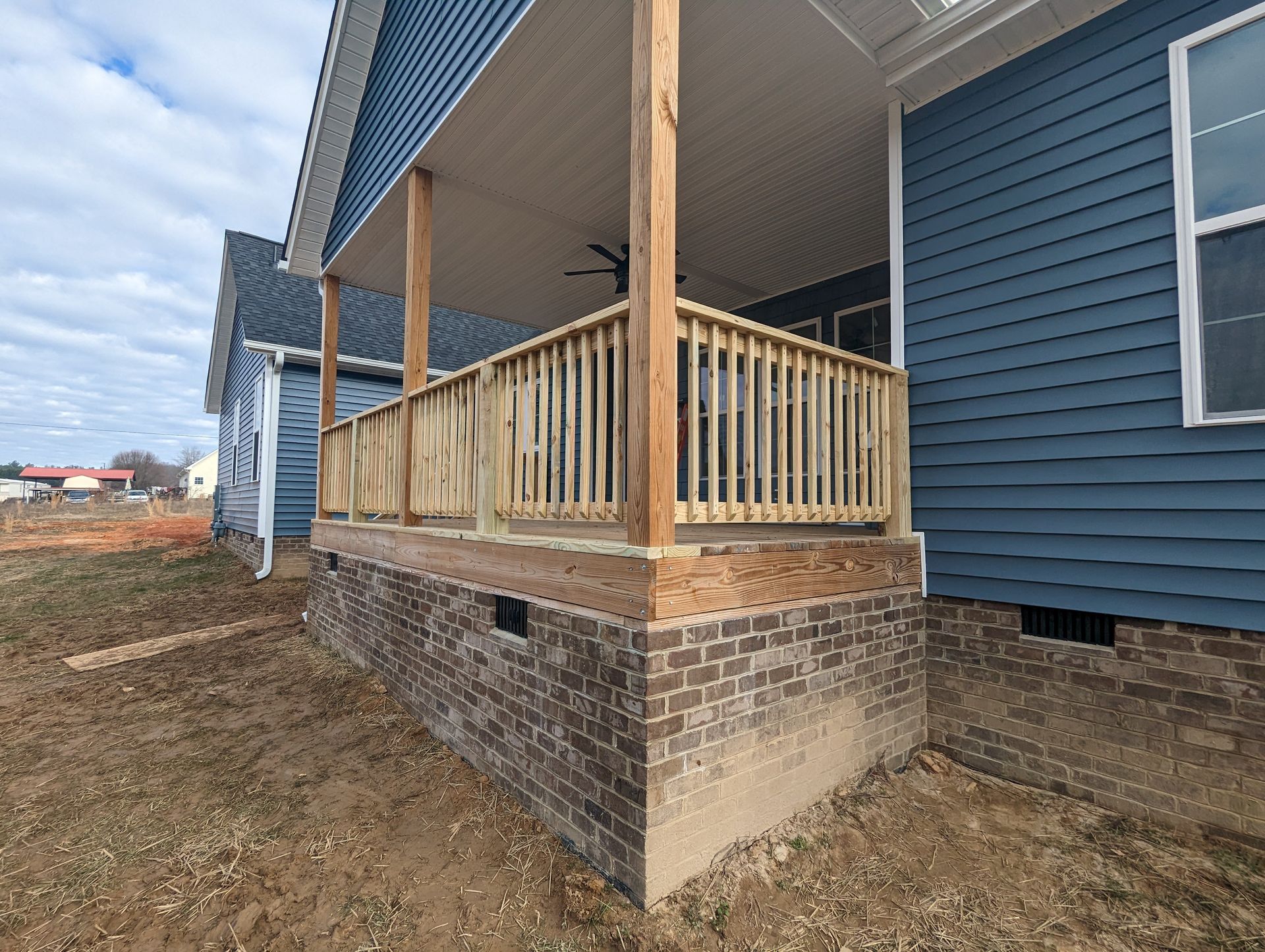 A newly constructed house with a front porch featuring wooden railings and brick foundation, set in a yard.