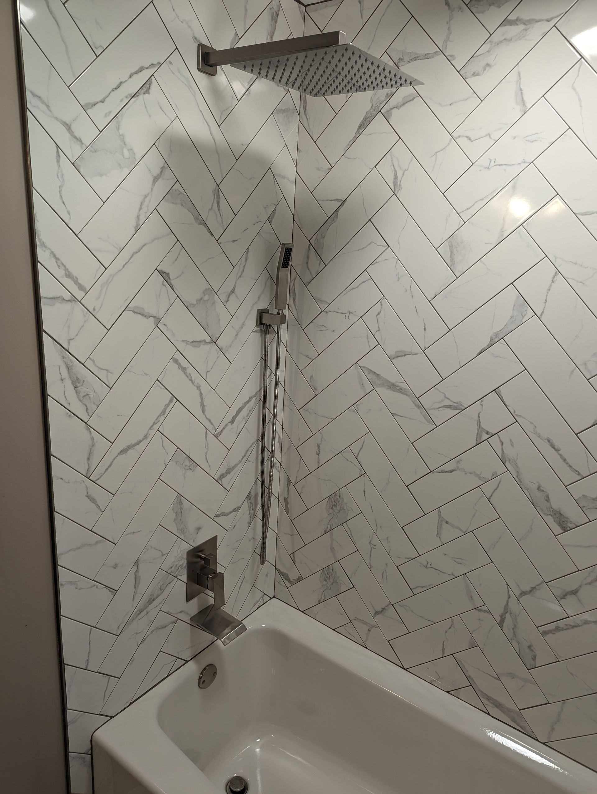A white and gray herringbone-tiled shower with a rain shower head, hand shower, and a white bathtub.