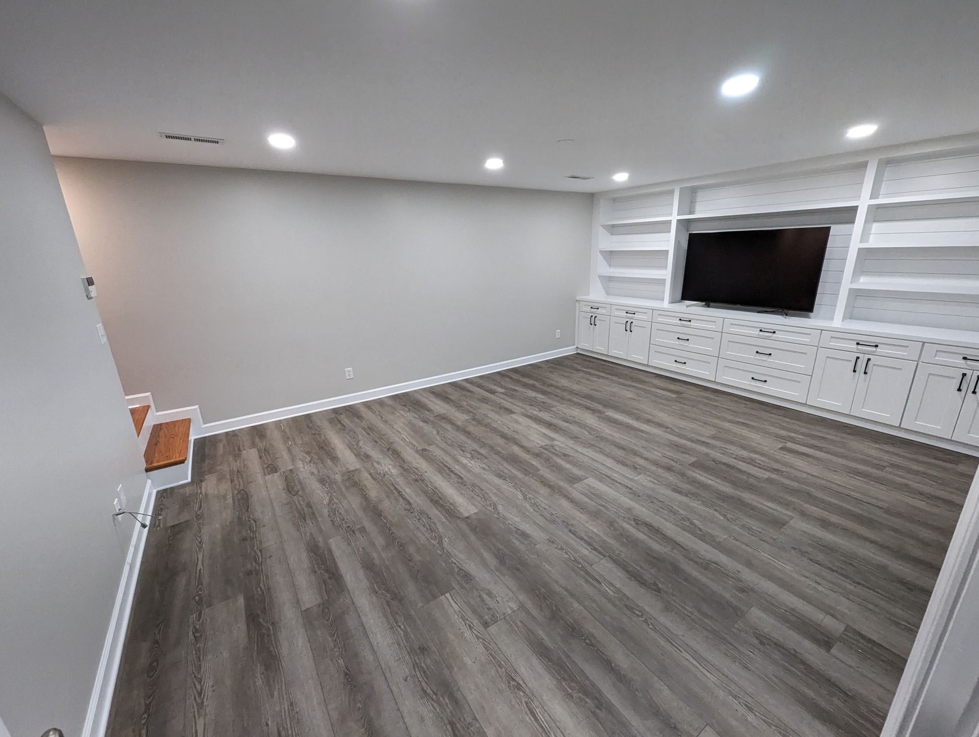 Empty living room with gray wood flooring and built-in white shelves around a large TV.