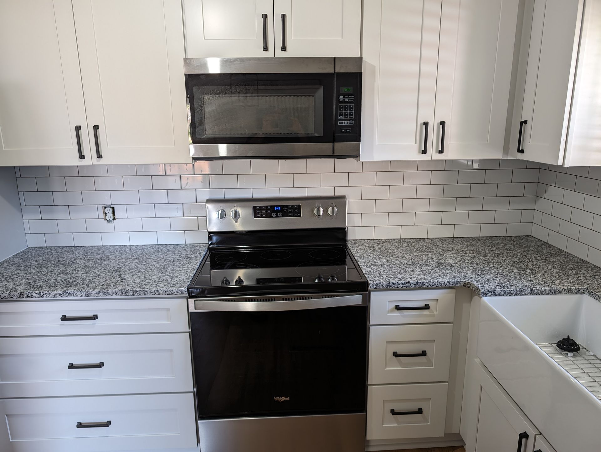 A modern white kitchen with black hardware. Includes white cabinets, a stainless steel microwave and stove, granite countertops, and white subway tile backsplash.