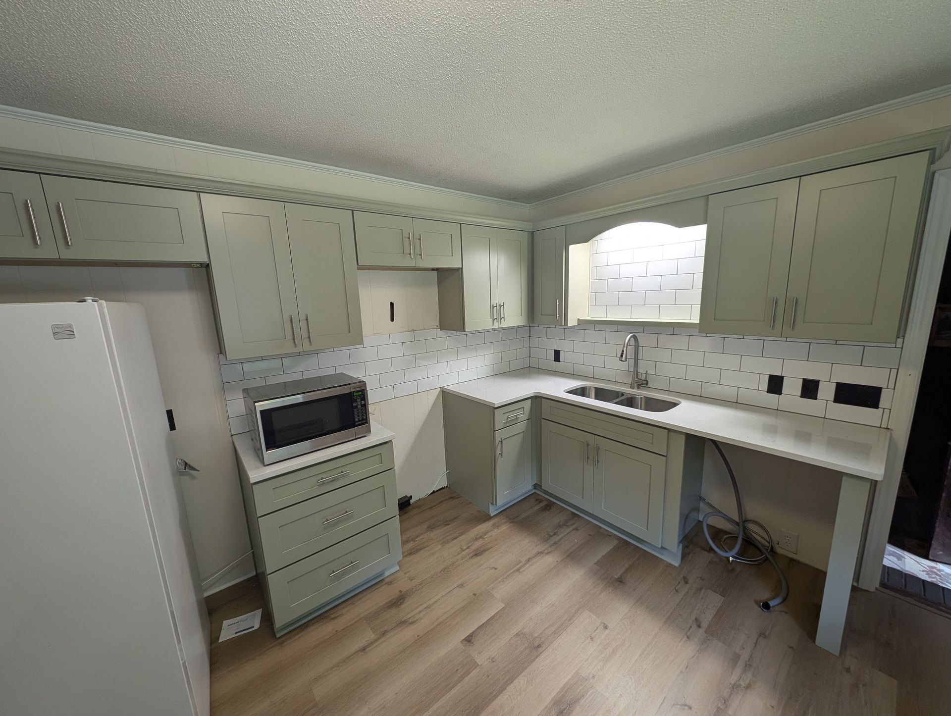 A small kitchen with light gray cabinets, a microwave, and a sink. The floor is light wood.