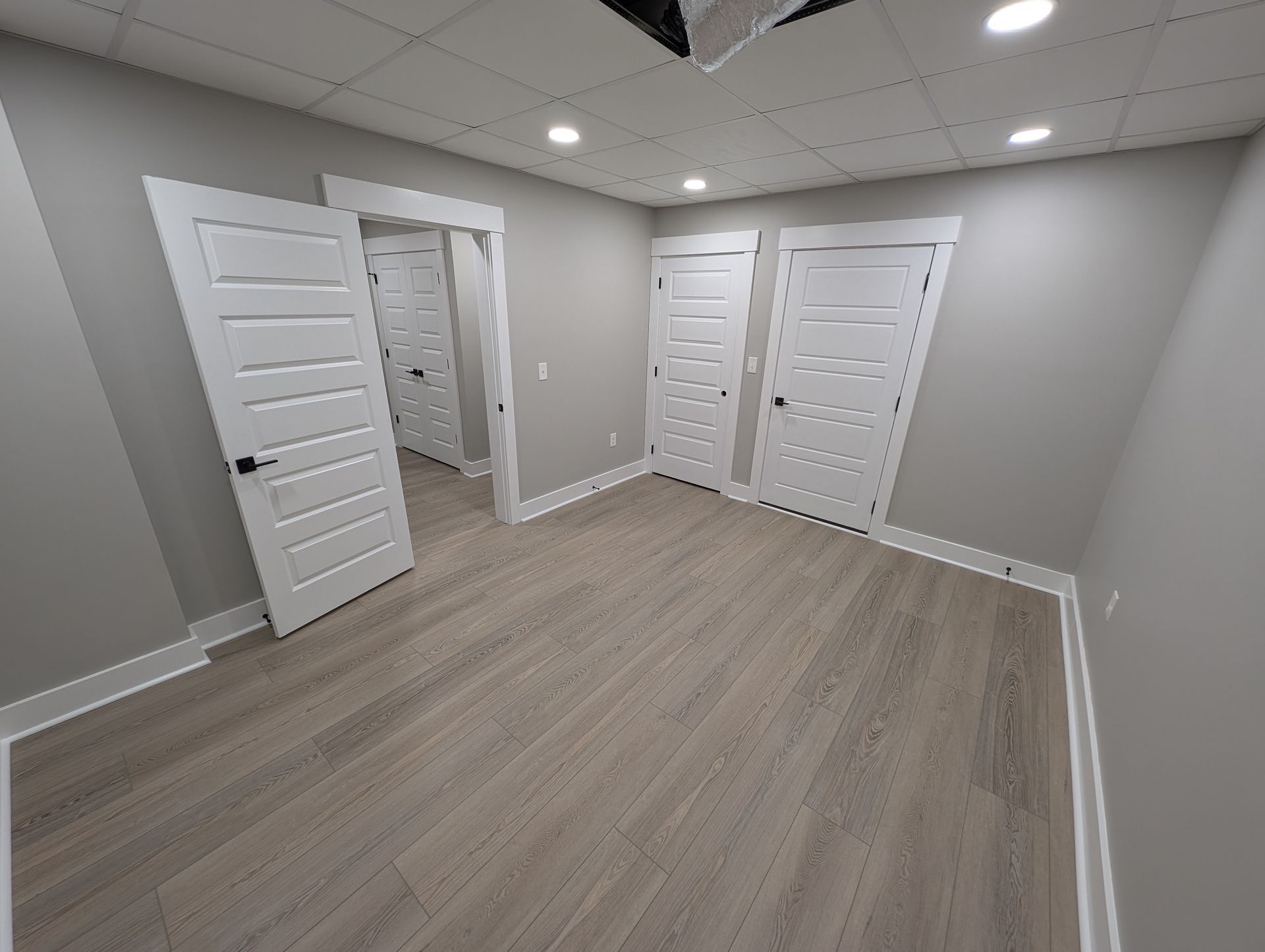 Empty room with light gray walls, light wood-look flooring, and several white doors with black hardware; recessed lighting.