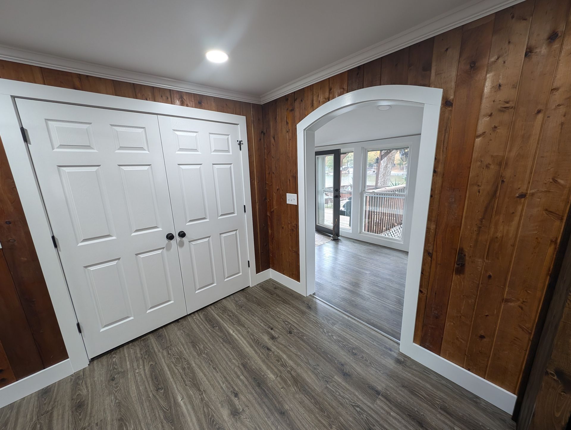 Interior with wood-paneled walls, white closet doors, and an arched doorway leading to a room with large windows.