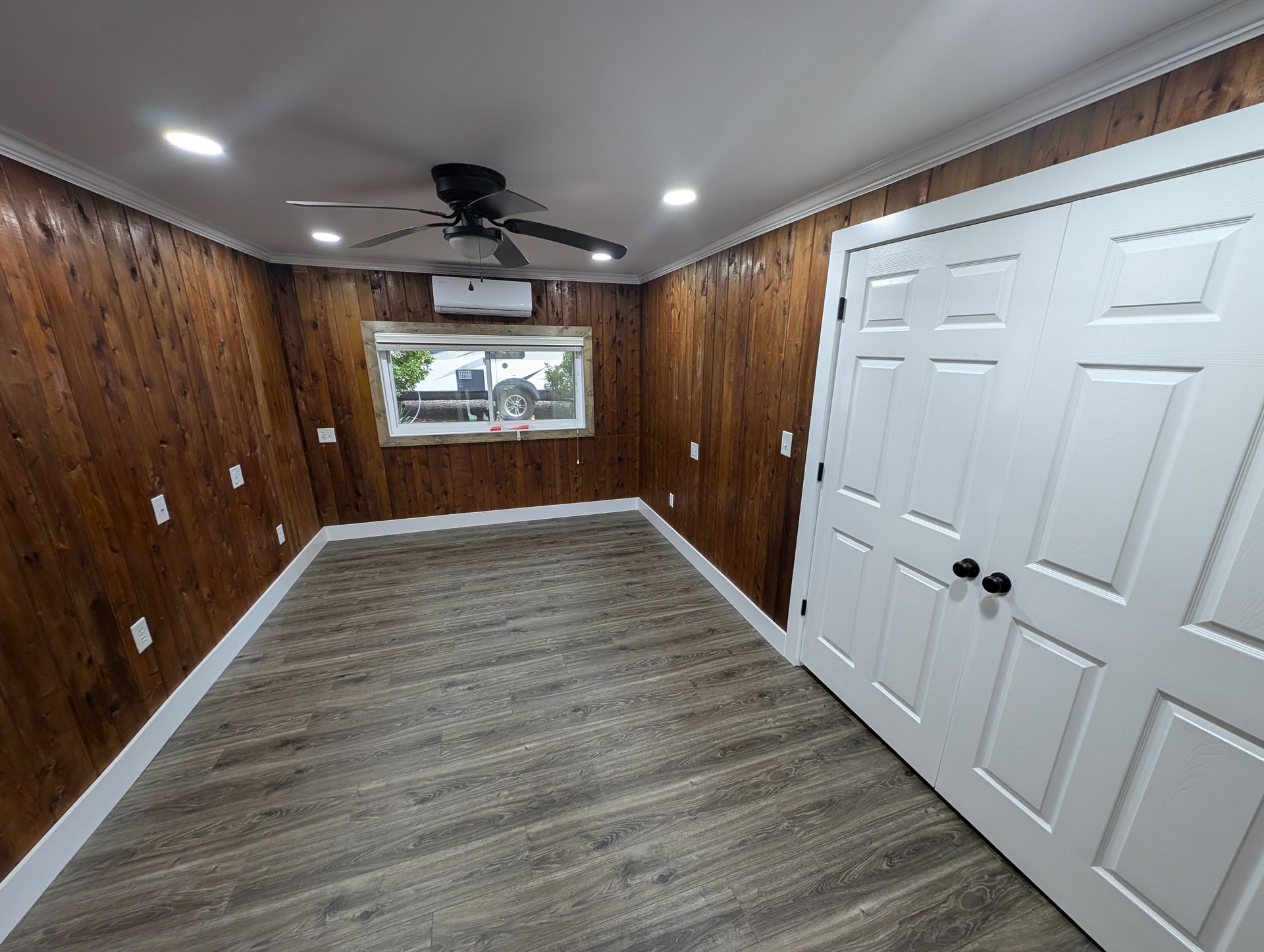 An empty room with dark wooden walls, a gray floor, and white trim. A window and a closed white door are also visible.