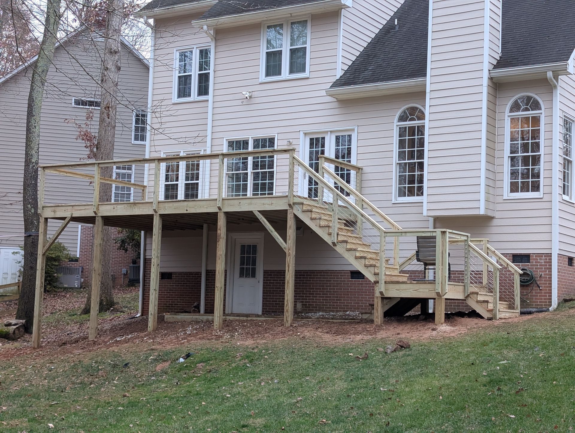 Back view of a two-story house with a wooden deck and stairs. Beige siding, brick foundation, and various windows are visible.