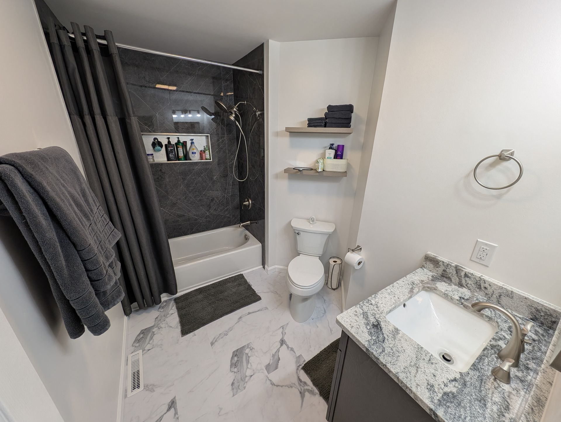 A modern bathroom with a marble floor and grey accents. Includes a shower, toilet, vanity, and shelving with towels and toiletries.