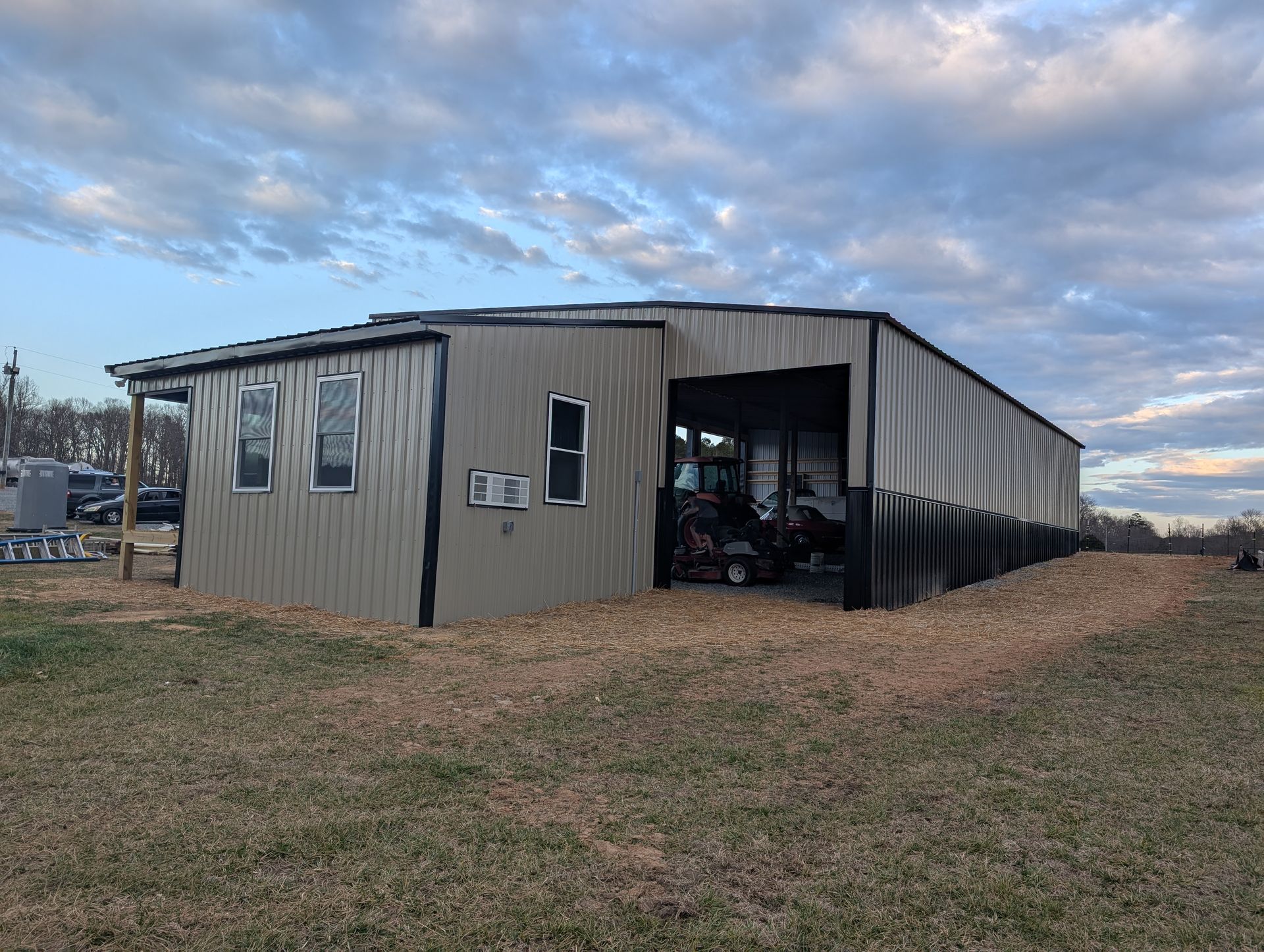 A tan metal shed with black accents, open on one side. It sits on a grassy area under a cloudy sky.