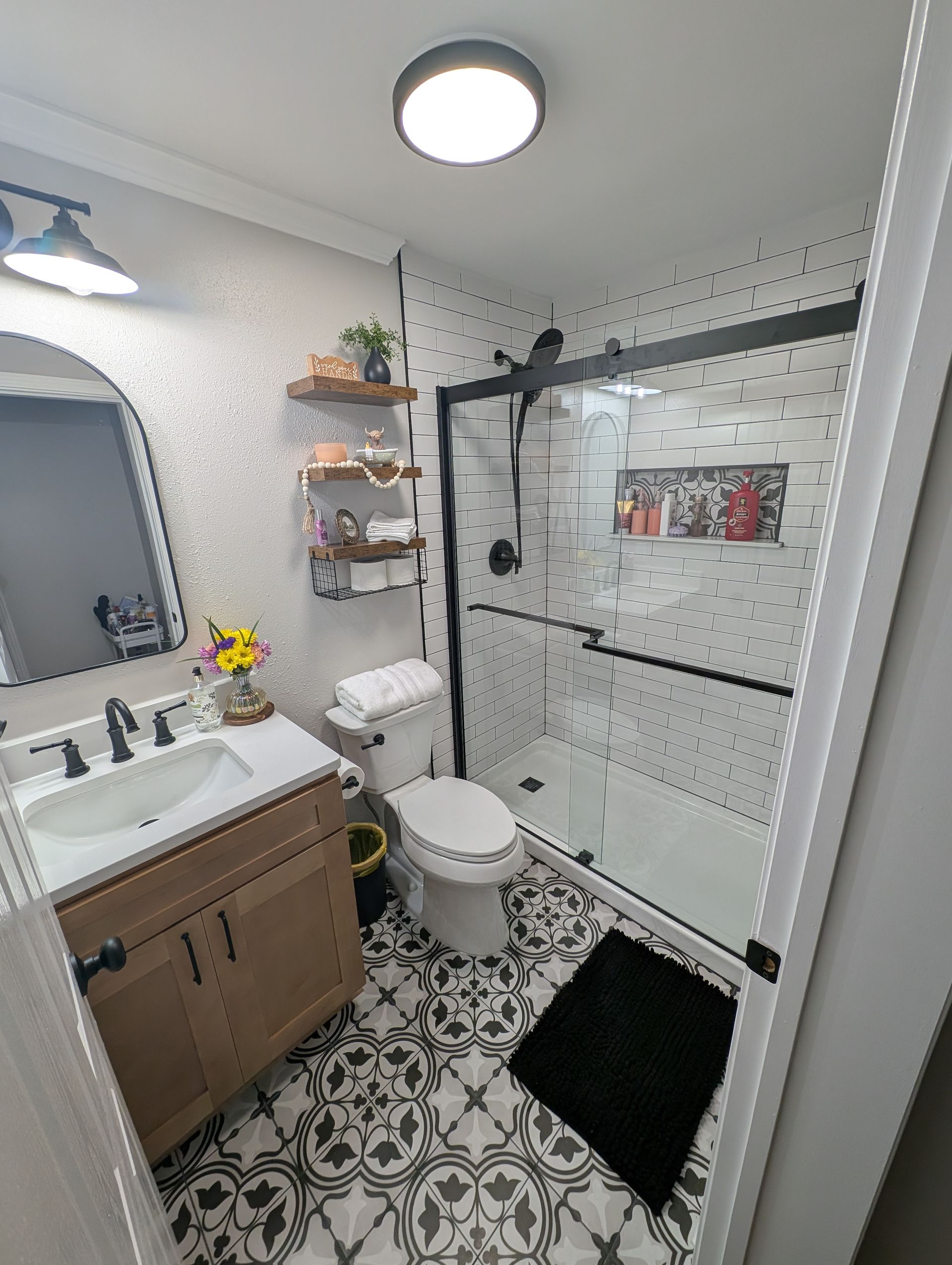 A newly renovated bathroom with patterned floor tiles, a black-framed shower, and a wooden vanity. The walls are white, some with black dot accents.