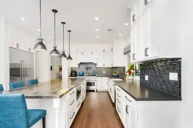Modern white kitchen with a granite island, pendant lighting, dark backsplash, and teal bar stools on hardwood floors.