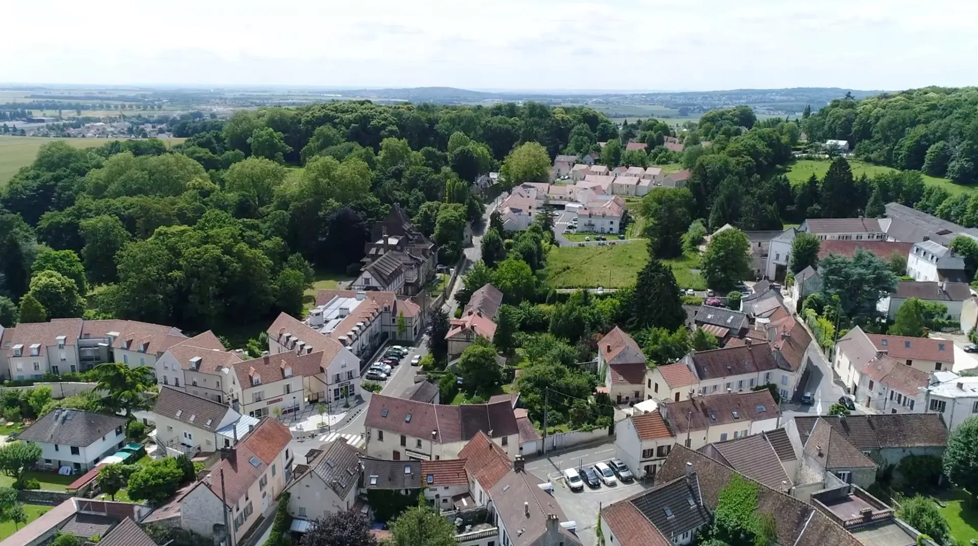 Vue aérienne d'un village aux bâtiments aux toits blancs, entouré d'arbres et de champs, par une journée ensoleillée.