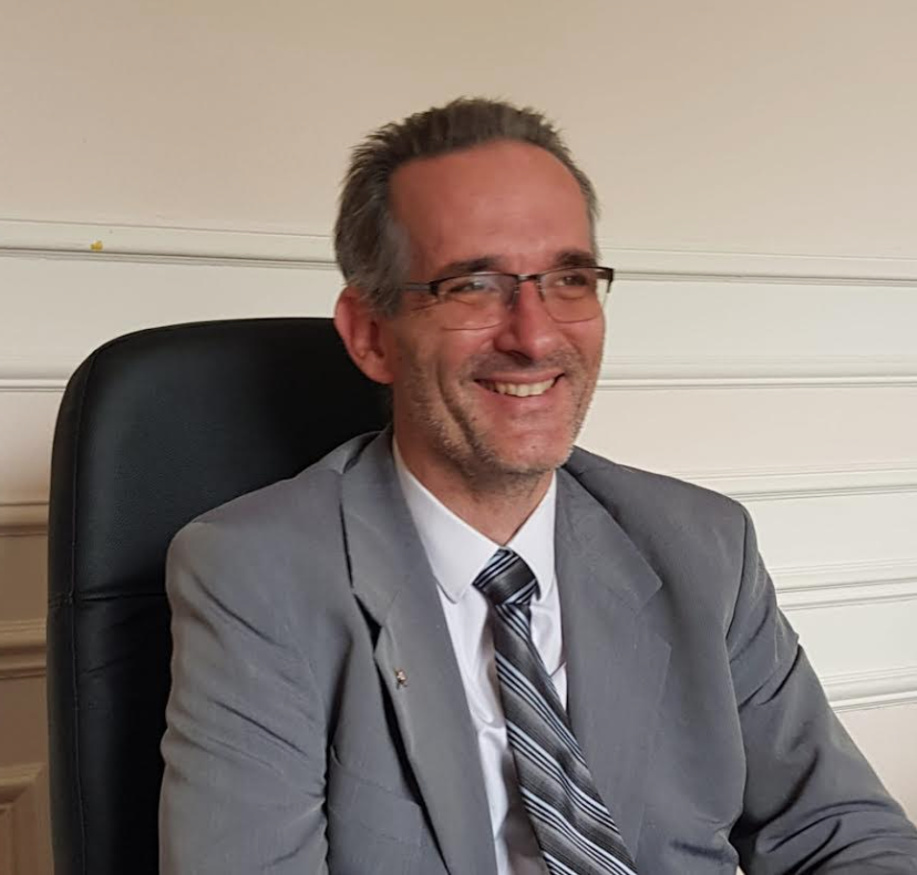 Un homme en costume gris sourit, portant des lunettes, assis à son bureau.