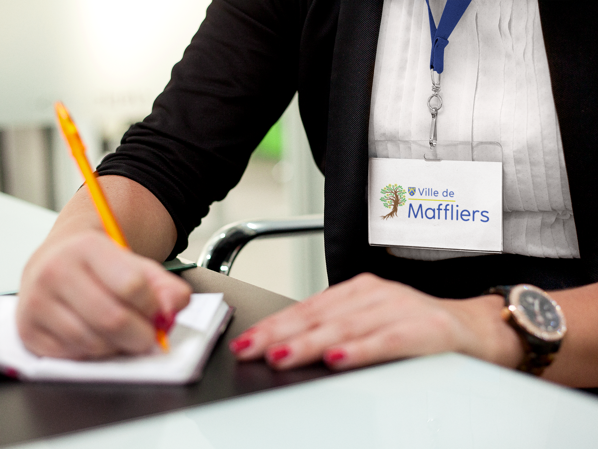 Femme écrivant dans un carnet, portant un badge avec le logo « Ville de Moffliers » et une montre.