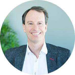 Portrait d'une personne souriante portant une chemise blanche à col et un blazer foncé texturé sur un fond bleu clair.