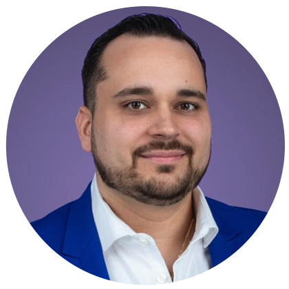 Une femme souriante aux cheveux blonds, vêtue d'un haut blanc à pois et de boucles d'oreilles en perles, se détache sur un fond de mur en pierre texturée.