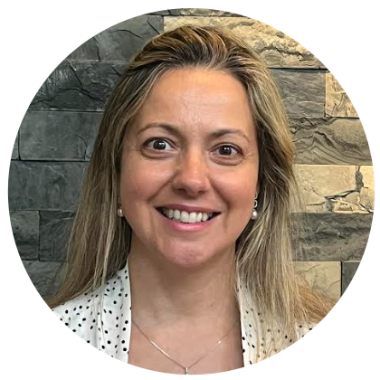 Une femme souriante aux cheveux blonds, vêtue d'un haut blanc à pois et de boucles d'oreilles en perles, se détache sur un fond de mur en pierre texturée.