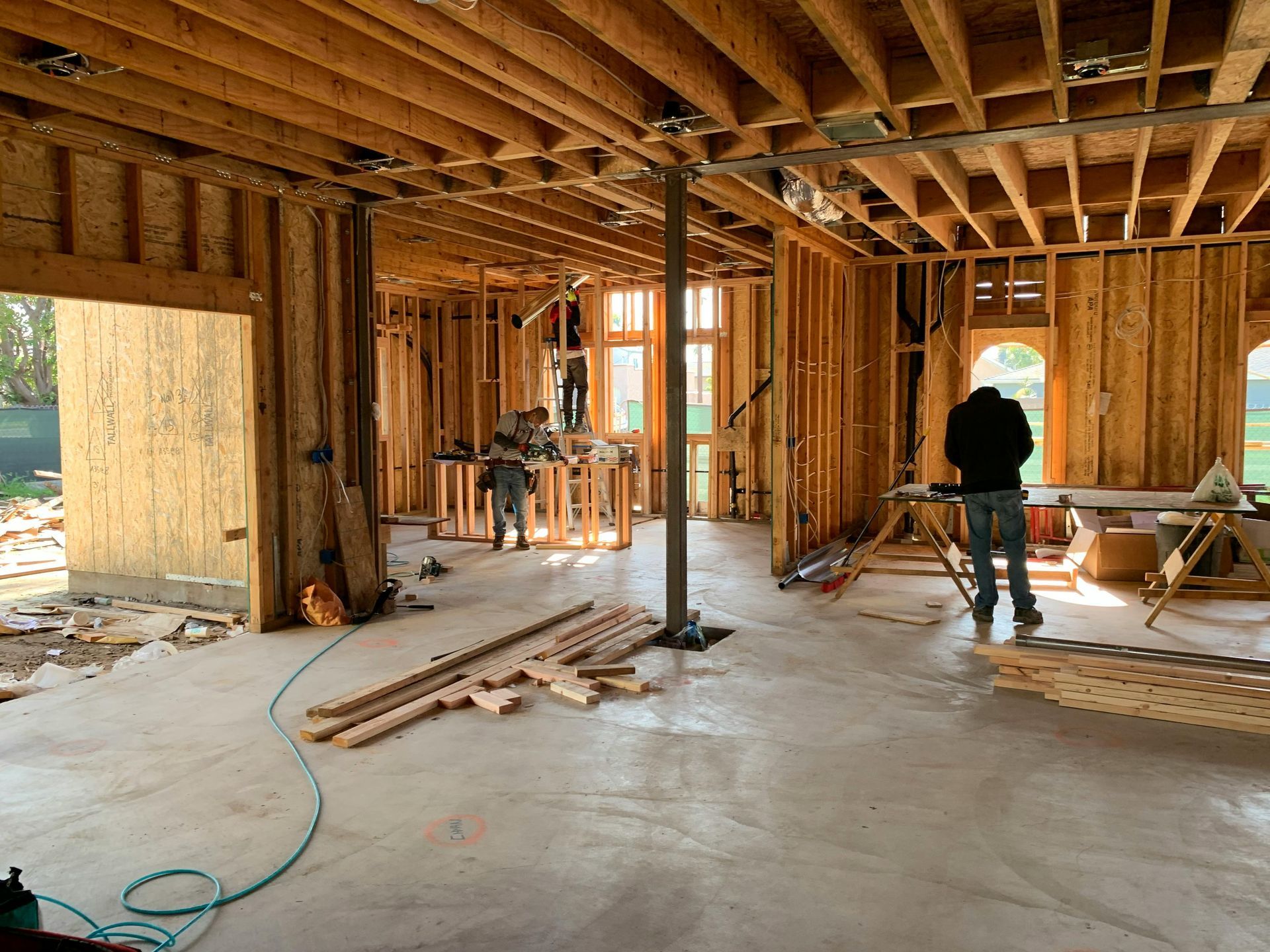 Intérieur d'un chantier de construction résidentielle avec charpente en bois apparente, sols en béton et deux ouvriers présents.