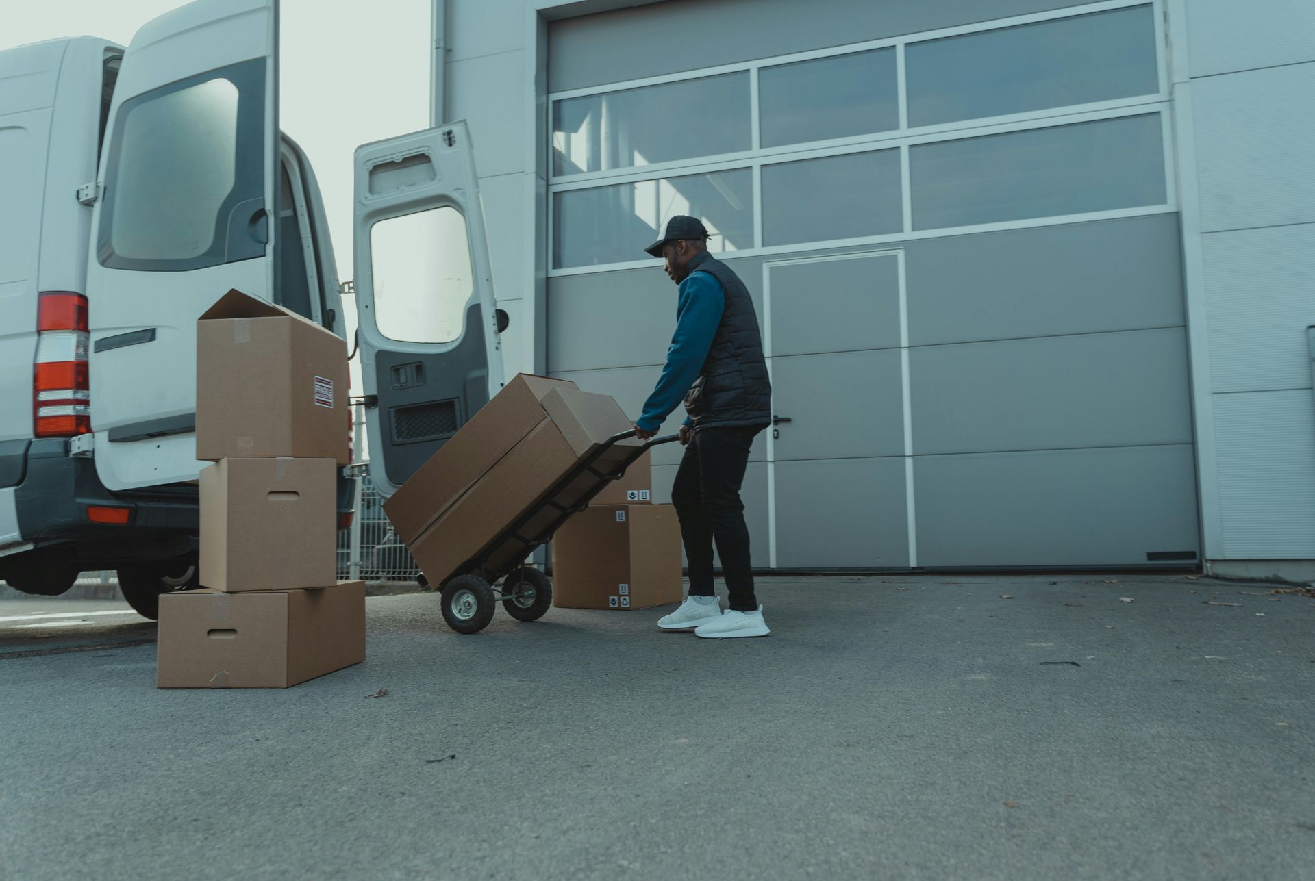 Un livreur utilise un diable pour déplacer des cartons de l'arrière d'une camionnette blanche stationnée devant un entrepôt.