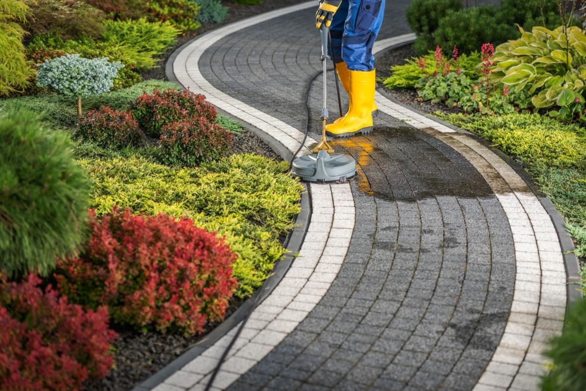 A man in yellow boots is cleaning a brick walkway in a garden.