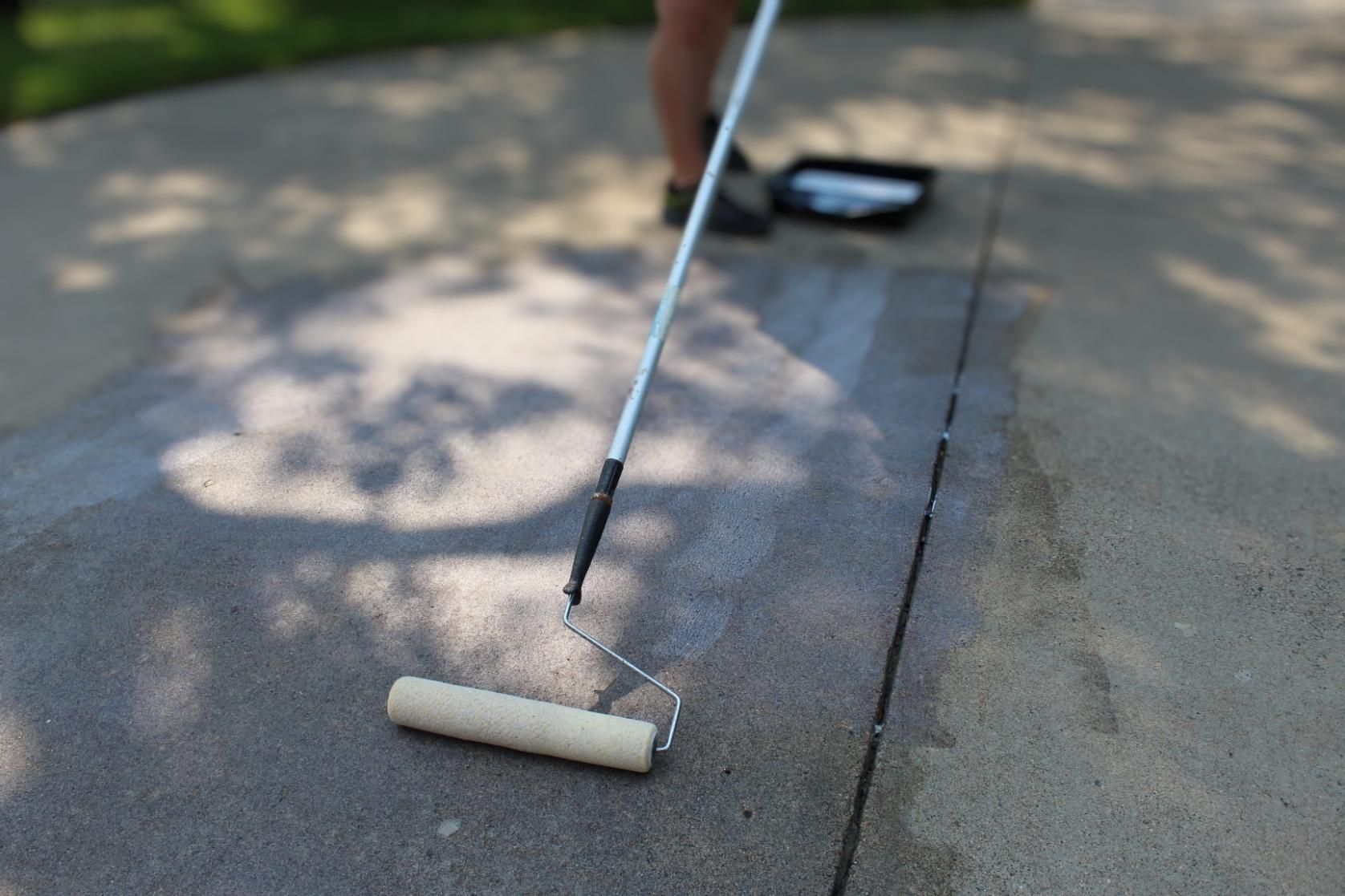 A person is using a paint roller to paint a concrete driveway.
