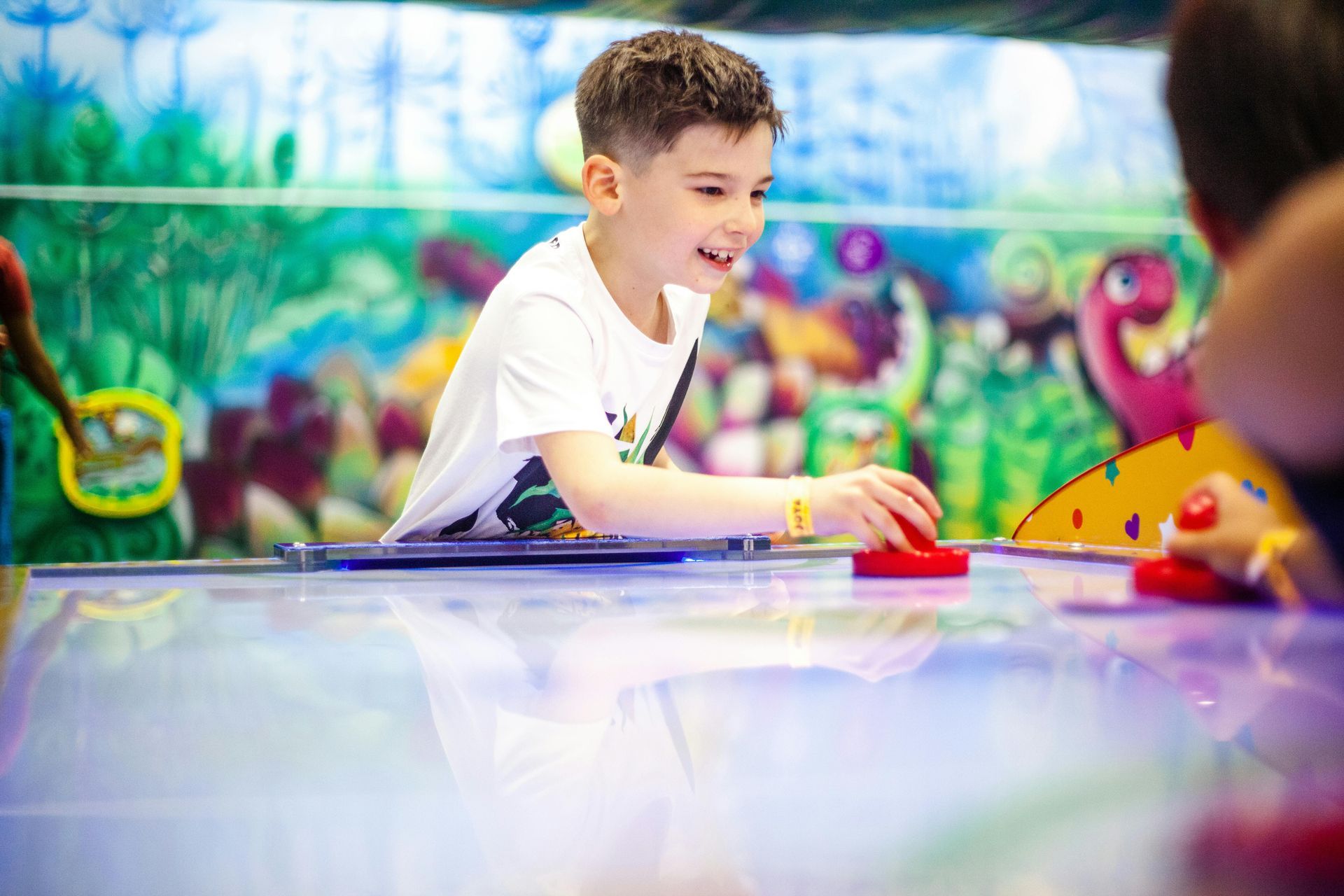 Boy smiles while playing air hockey at a brightly colored arcade game with a jungle backdrop.
