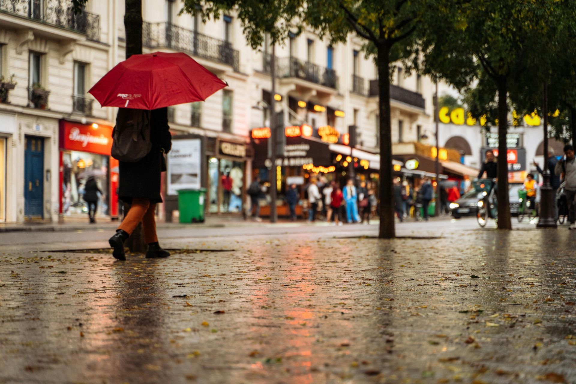 Une personne marche dans une rue mouillée, sous un parapluie rouge, avec des devantures de magasins et des piétons flous en arrière-plan.
