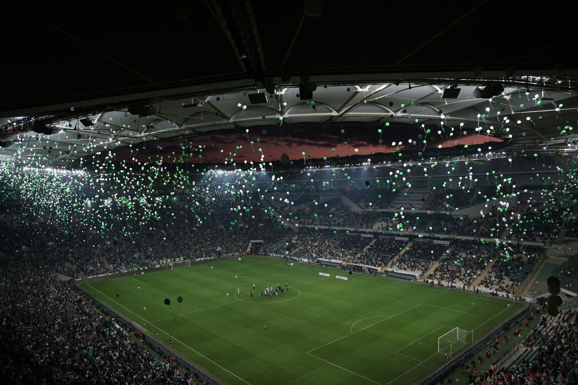 Un match de football nocturne dans un stade illuminé de fumigènes verts ; les joueurs évoluent sur le terrain vert.