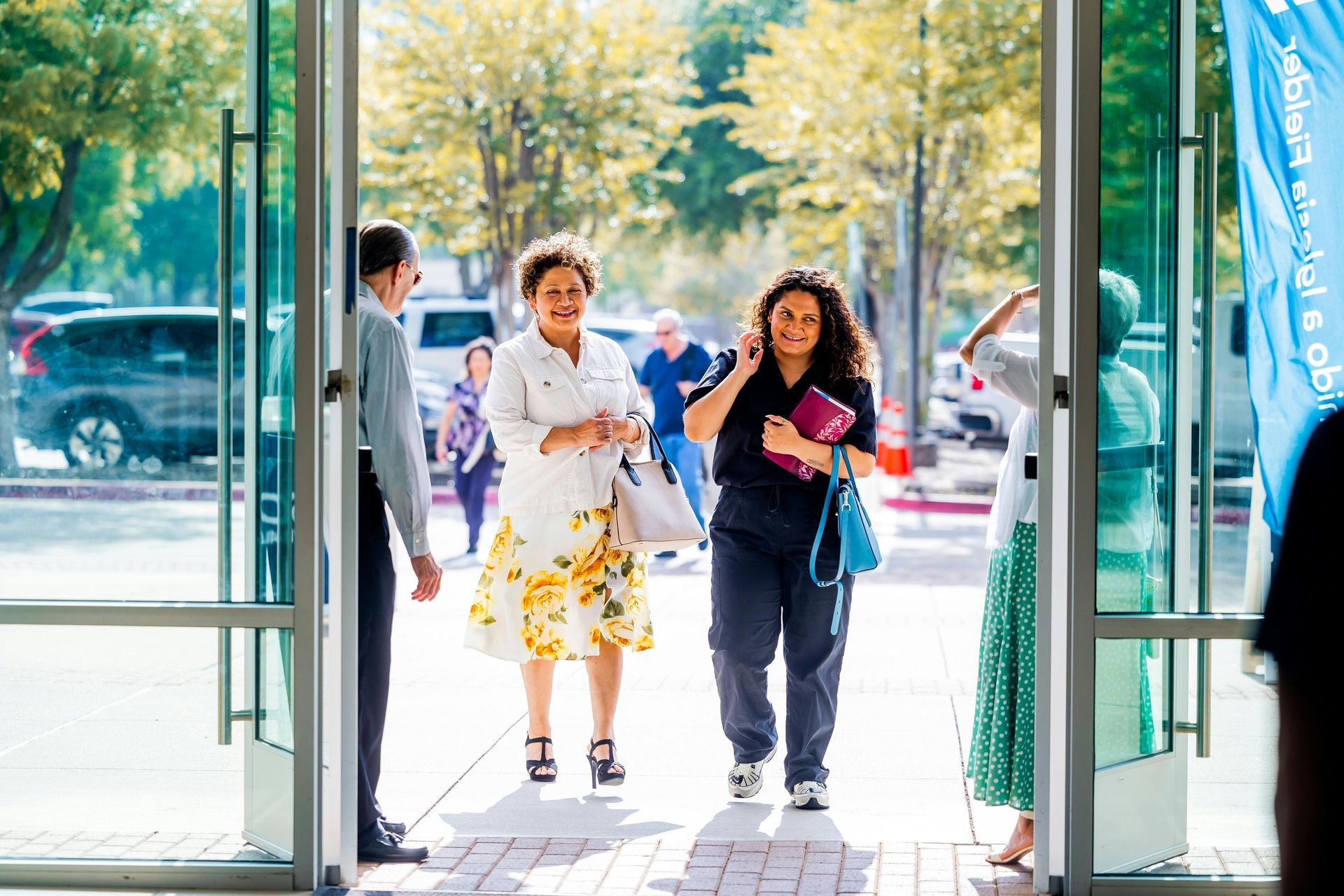 Deux femmes sortent d'un immeuble par des portes vitrées et s'avancent dans la lumière du soleil.