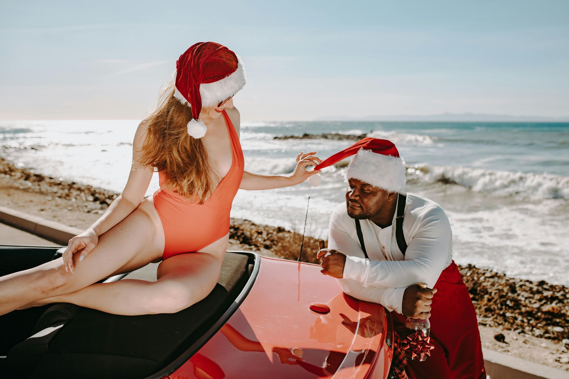Une femme en maillot de bain et bonnet de Père Noël tire sur le bonnet d'un homme déguisé en Père Noël, appuyé contre une voiture rouge sur la plage.