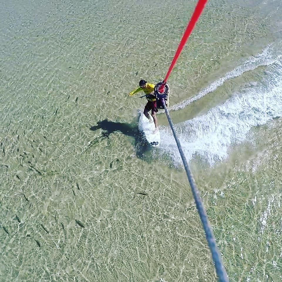 Kiteboarder su una tavola bianca in acque basse e limpide vicino a una spiaggia. Sono visibili le linee rosse dell'aquilone, con una maglietta gialla.