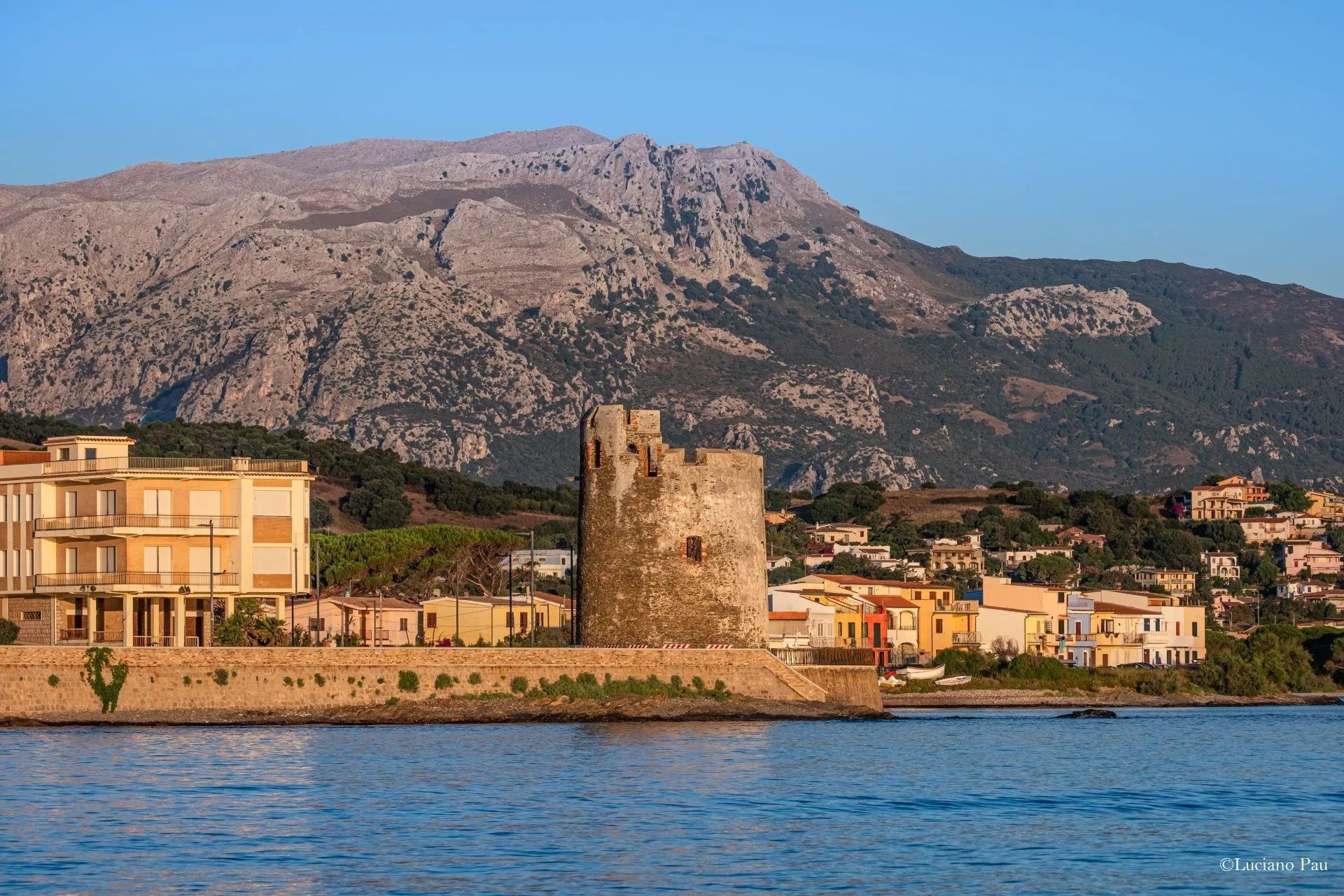 Vista costiera con una torre in pietra, edifici e una montagna contro il cielo azzurro.