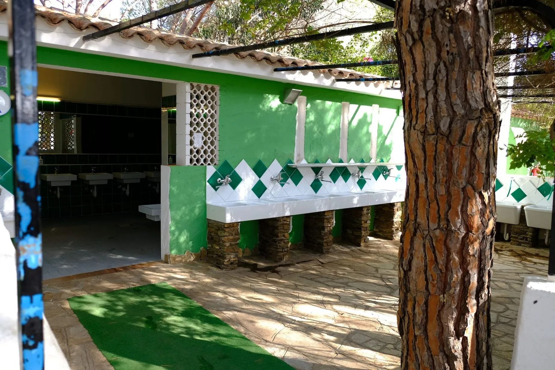 Bagno esterno verde e bianco con più lavandini. In primo piano un tronco d'albero.