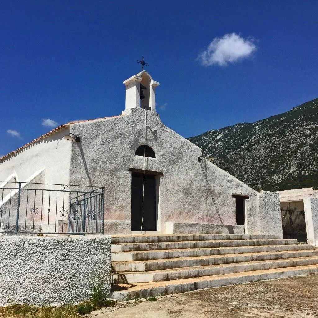 Chiesa bianca con gradini, croce e campanile sullo sfondo del cielo azzurro e delle montagne.