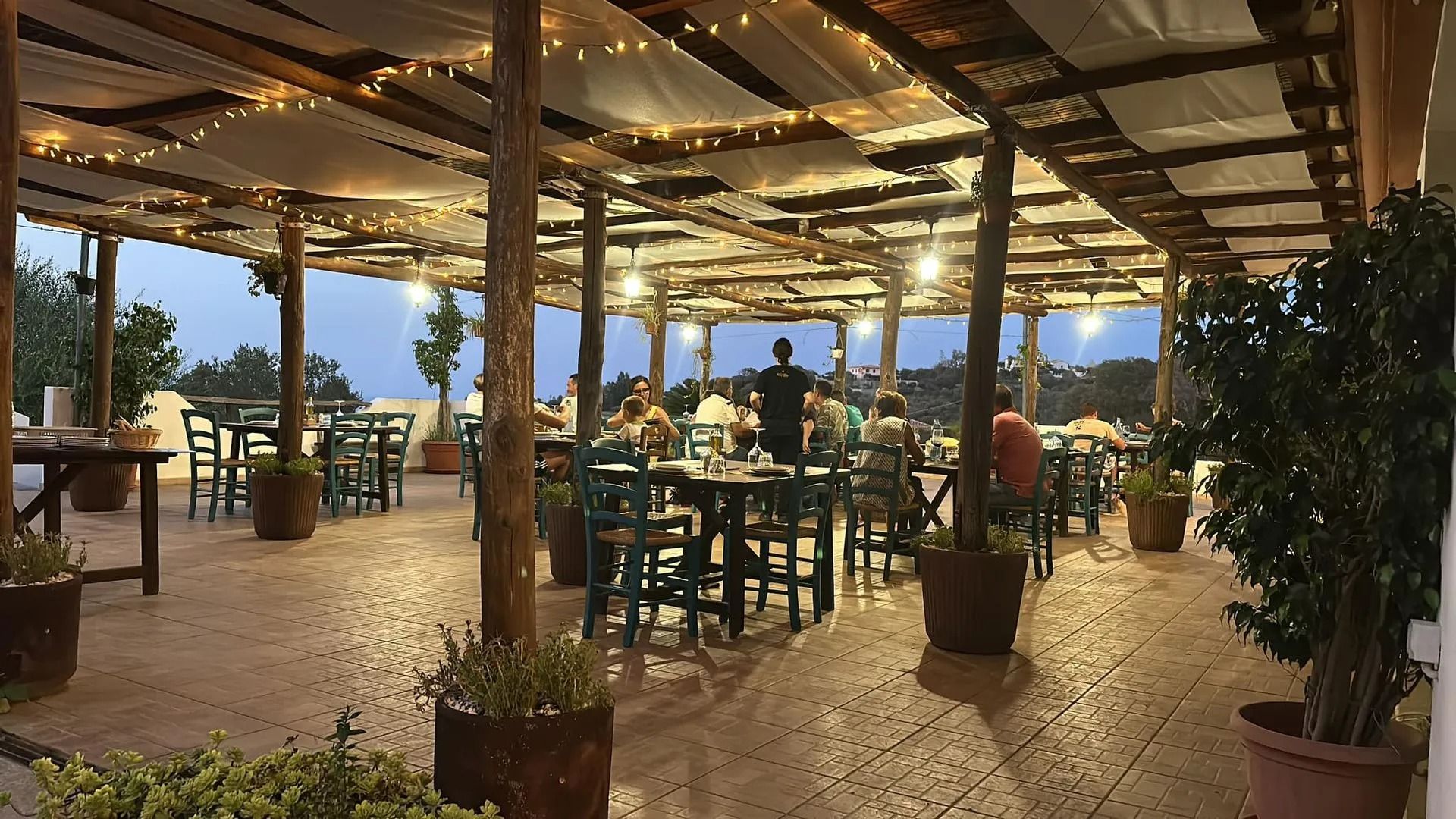 Ristorante all'aperto sotto una tettoia in legno con tavoli, sedie e lucine. Persone che cenano.