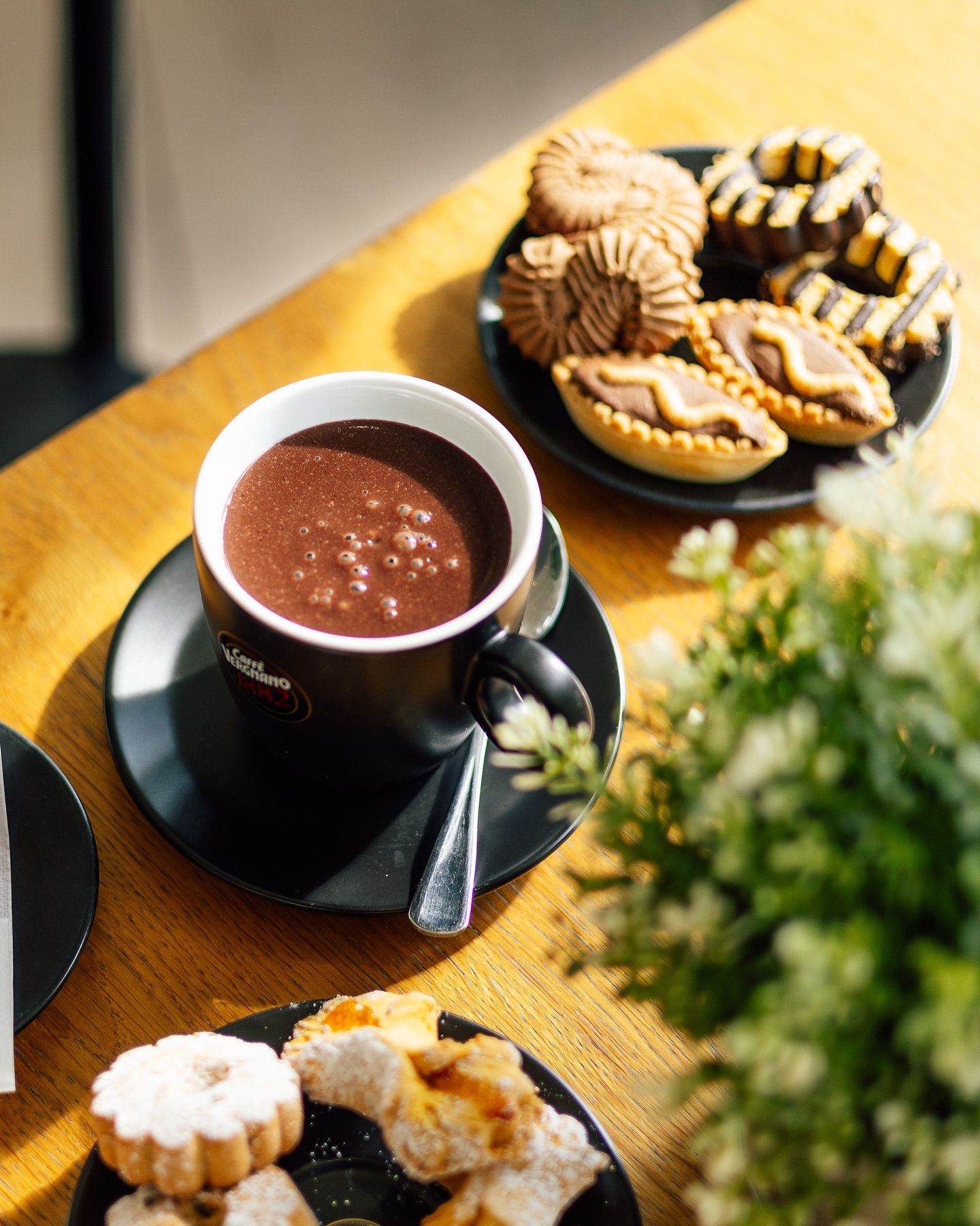 Cioccolata calda in una tazza nera con biscotti su un tavolo di legno, luce del sole.