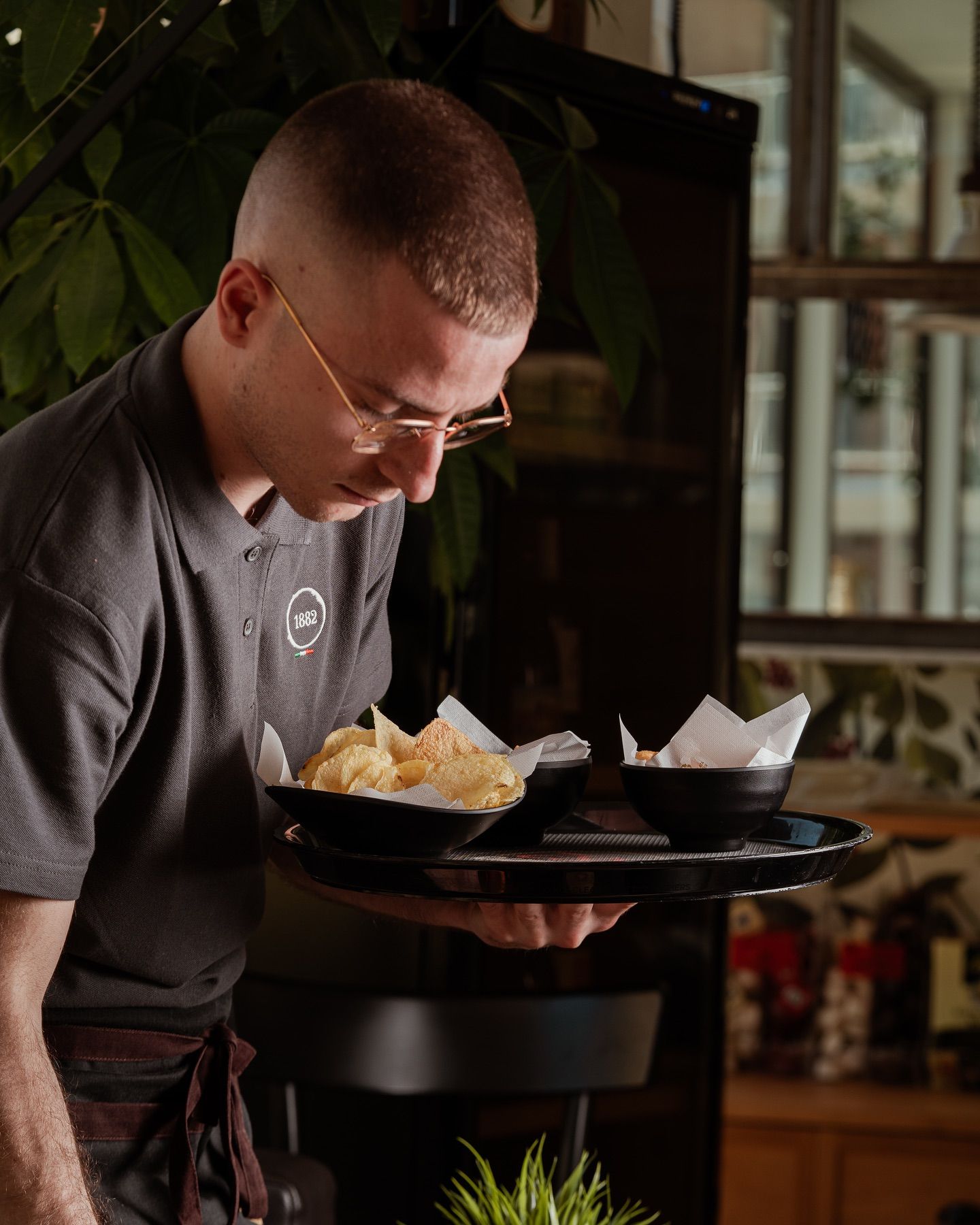 Cameriere che tiene un vassoio con del cibo, guardando in basso. Vassoio nero, camicia grigia, occhiali, ambiente interno.