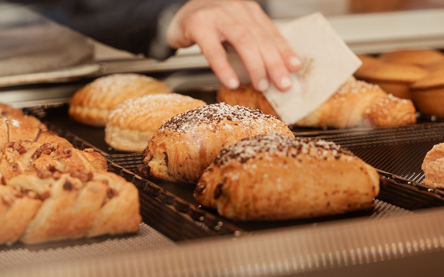 Pasticcini esposti in una vetrina di panetteria. Mano che appoggia un foglio di carta vicino a un croissant.