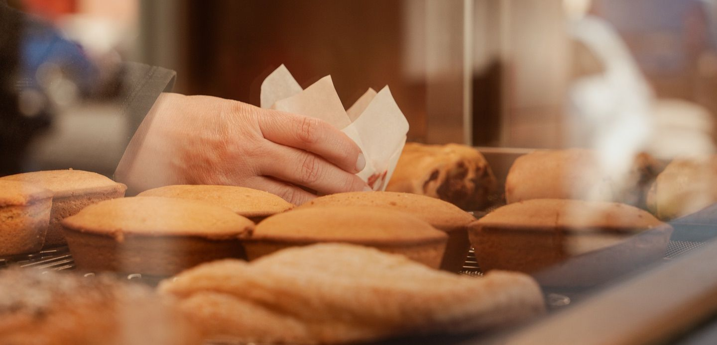 Una mano che tiene un involucro di carta sopra prodotti da forno, probabilmente muffin, dietro il vetro di una panetteria.