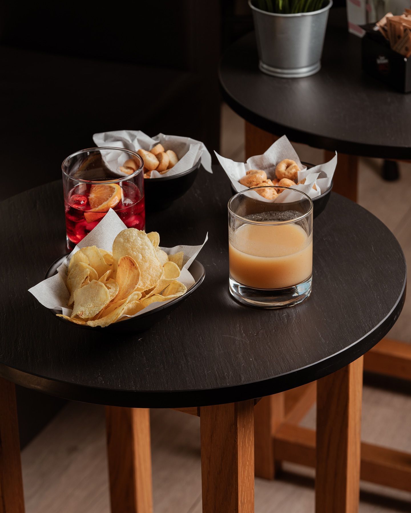 Bevande e snack su piccoli tavoli neri, tra cui patatine, biscotti e cocktail in bicchieri.
