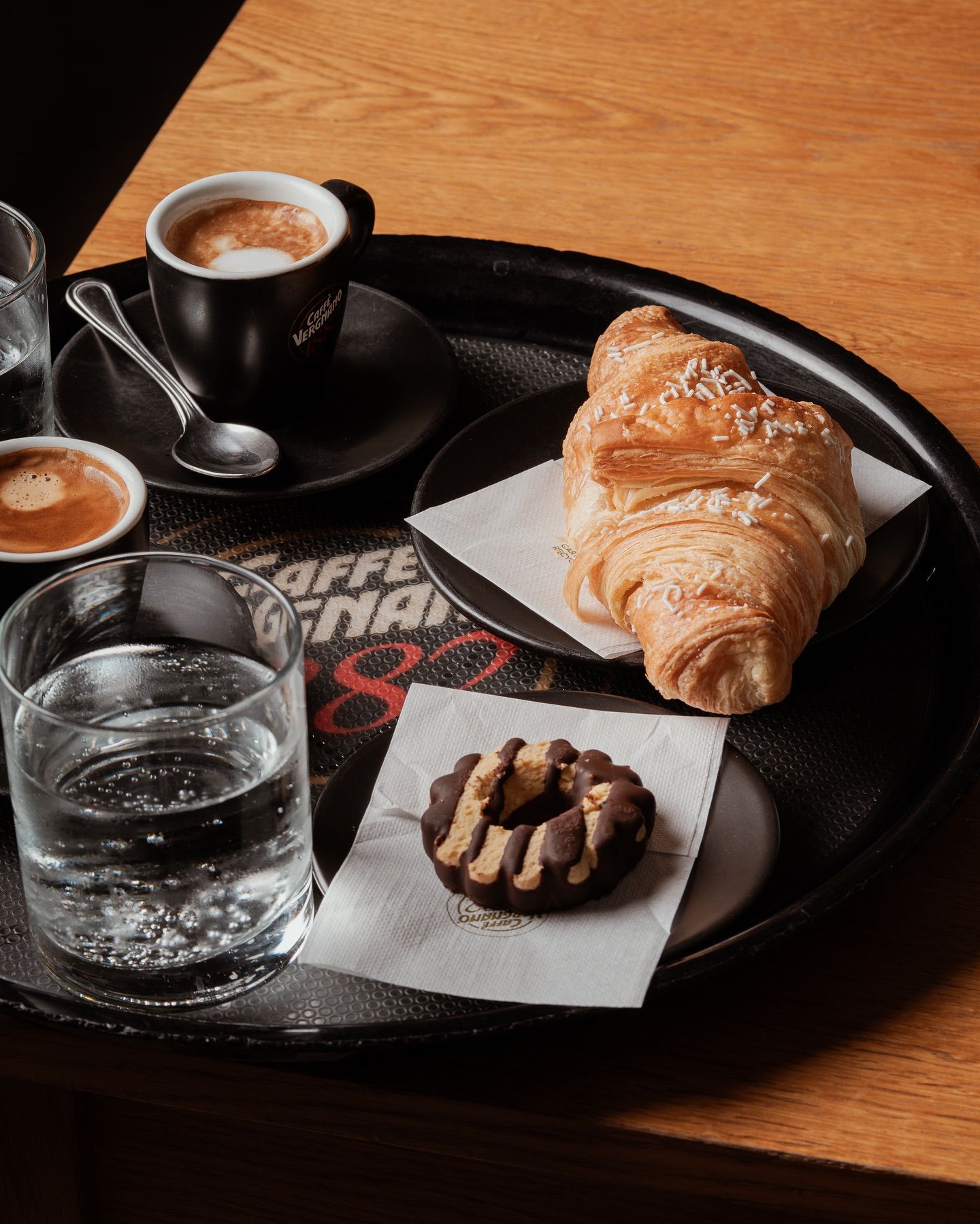 Vassoio con caffè, croissant, biscotti e bicchieri d'acqua su un tavolo di legno.